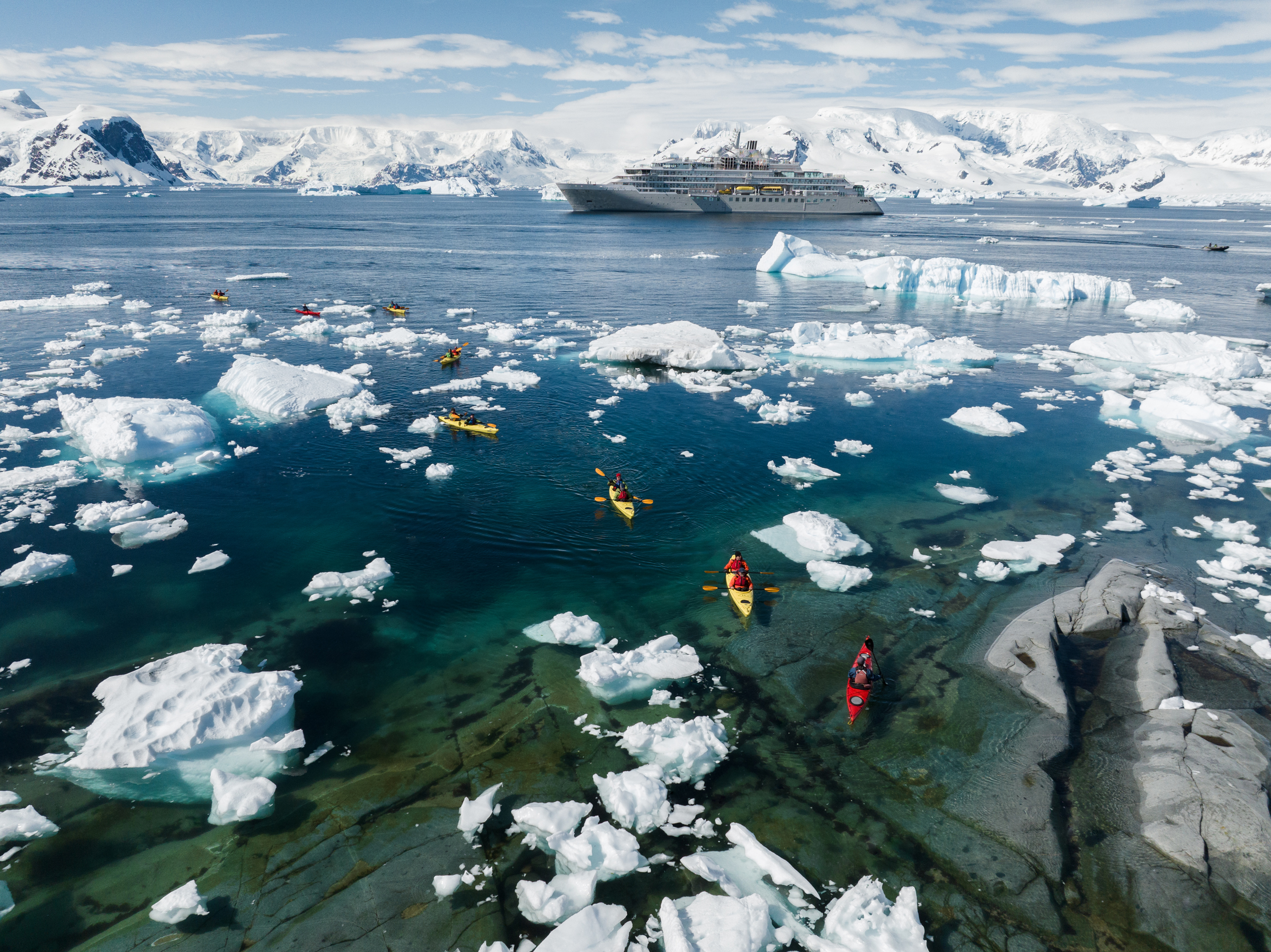 Kayakers in colorful kayaks paddling among icebergs with a cruise ship and snow-covered mountains