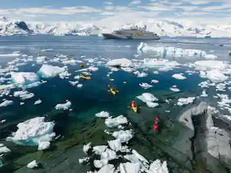 Kayakers in colorful kayaks paddling among icebergs with a cruise ship and snow-covered mountains