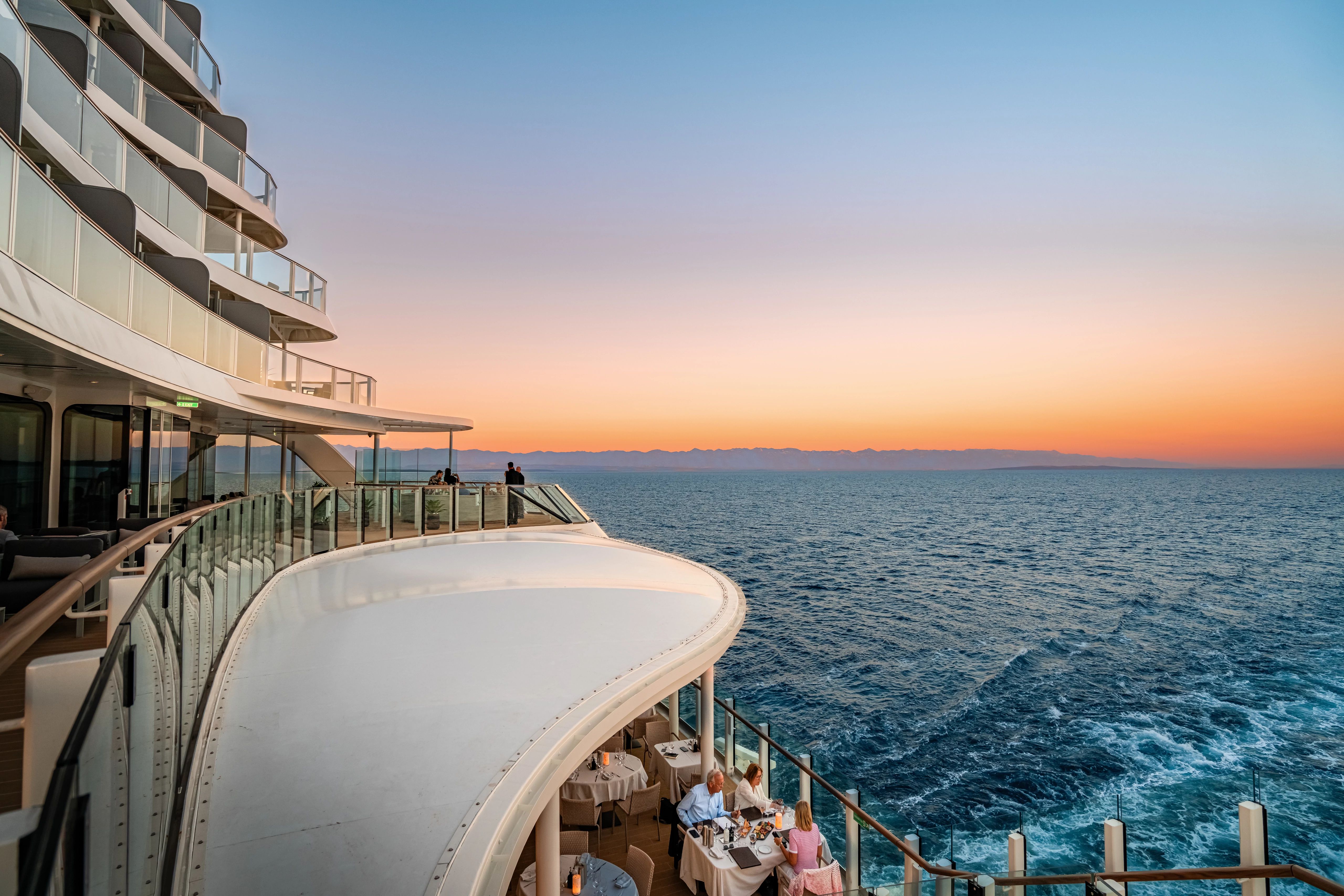 Curved cruise ship balconies with glass railings and diners overlooking ocean at sunset