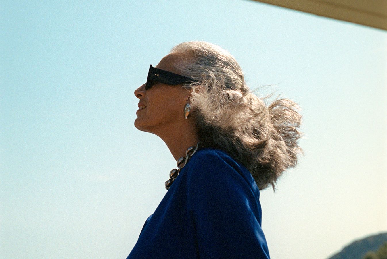 Older woman in sunglasses and blue jacket, silver hair blowing, necklace and earrings, looking up