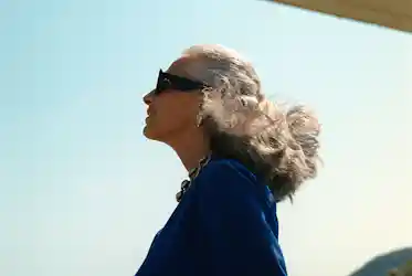 Older woman in sunglasses and blue jacket, silver hair blowing, necklace and earrings, looking up