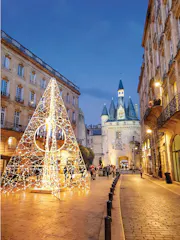 Christmas light pyramid on plaza beside historic stone gate with conical towers at dusk