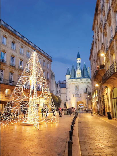 Christmas light pyramid on plaza beside historic stone gate with conical towers at dusk