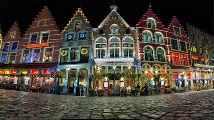 Nighttime row of gabled Flemish buildings lit with holiday lights, wreaths and outdoor cafe seating