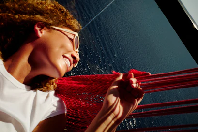 Smiling person in sunglasses reclining in red hammock over sparkling blue water