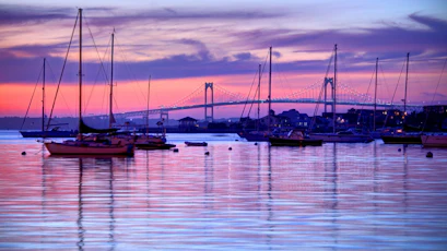 Sunset harbor with anchored sailboats, tall masts, reflections and a lit suspension bridge