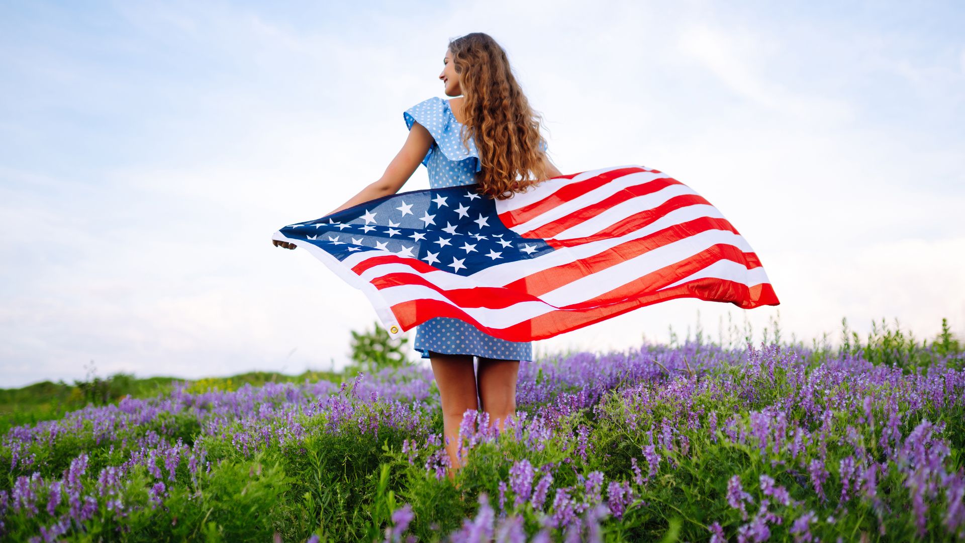 Woman with long curly hair in blue polka-dot dress in purple wildflowers holding American flag