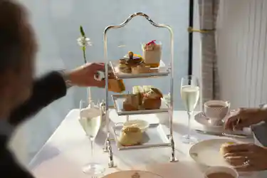 Three-tier afternoon tea stand with sandwiches, scones, pastries, two champagne flutes and hands.
