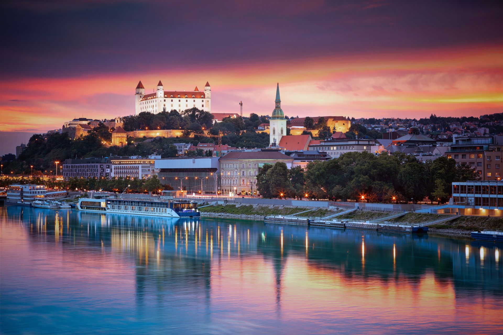 Bratislava Castle and spired cathedral above the Danube at sunset, boats reflected