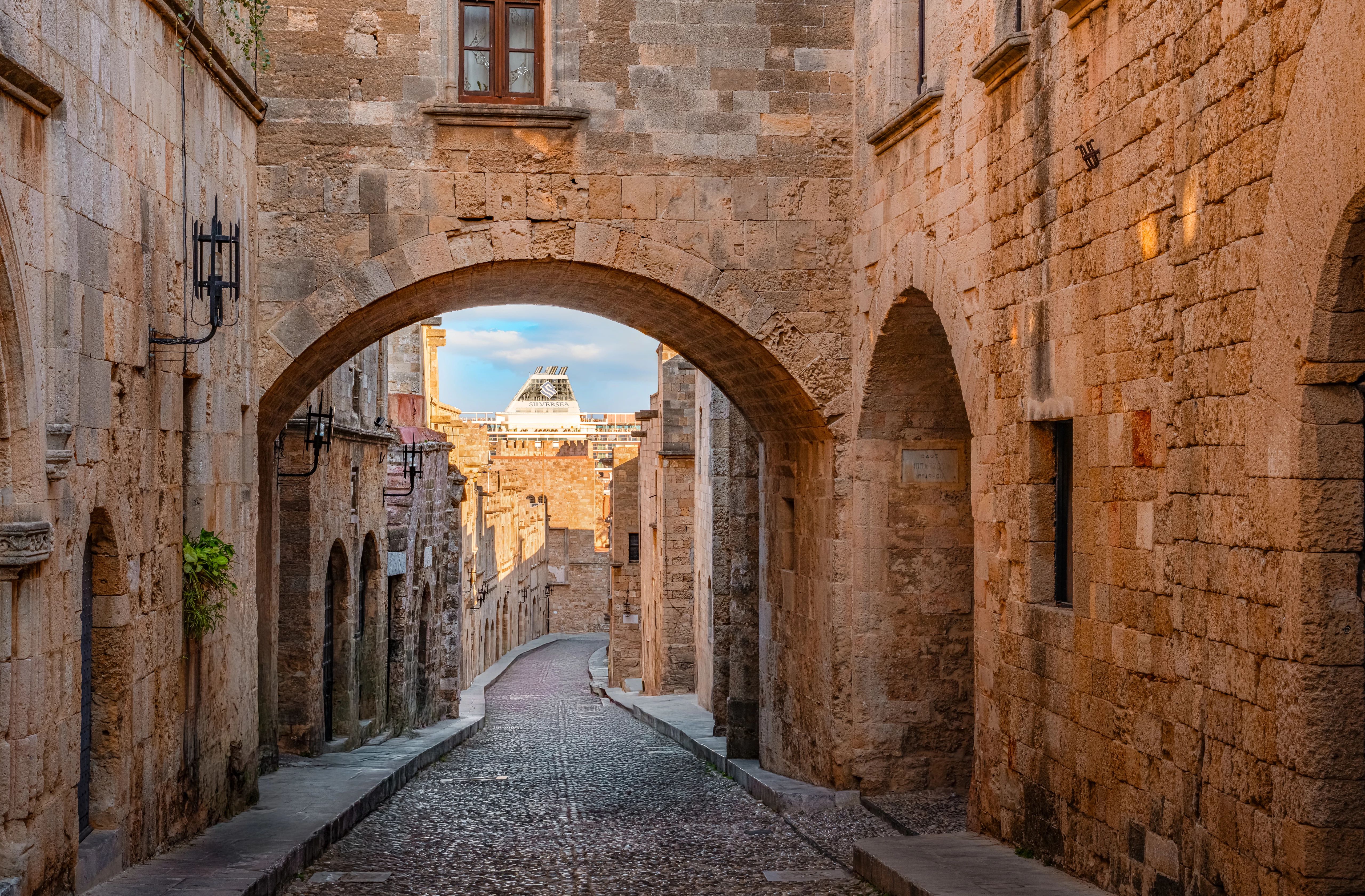 Narrow cobbled medieval street framed by stone arches and a distant cruise ship funnel