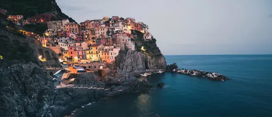 Cliffside Mediterranean village at dusk with colorful houses, lit harbor and boats