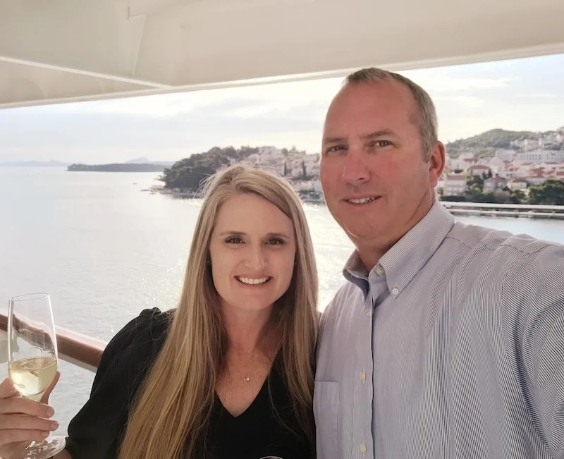 Two people taking a selfie on a cruise balcony with wine glass, coastal town and sea behind