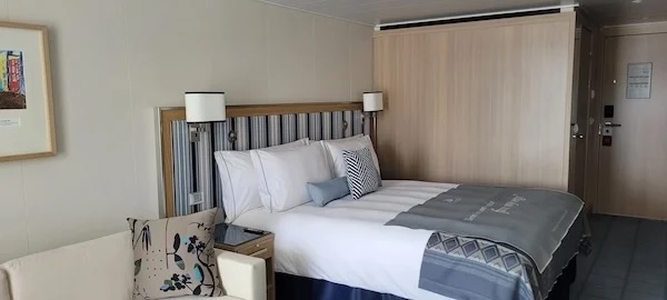 Double bed with white linens, throw and pillows, bedside lamps, sofa with patterned cushion in cabin