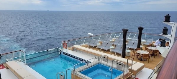 Cruise ship aft deck with swimming pool, hot tub, sun loungers, tables and open ocean horizon