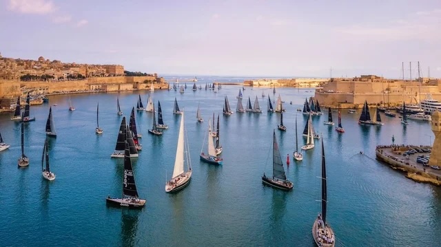 Numerous sailboats in a regatta crossing a wide harbor flanked by historic stone fortifications