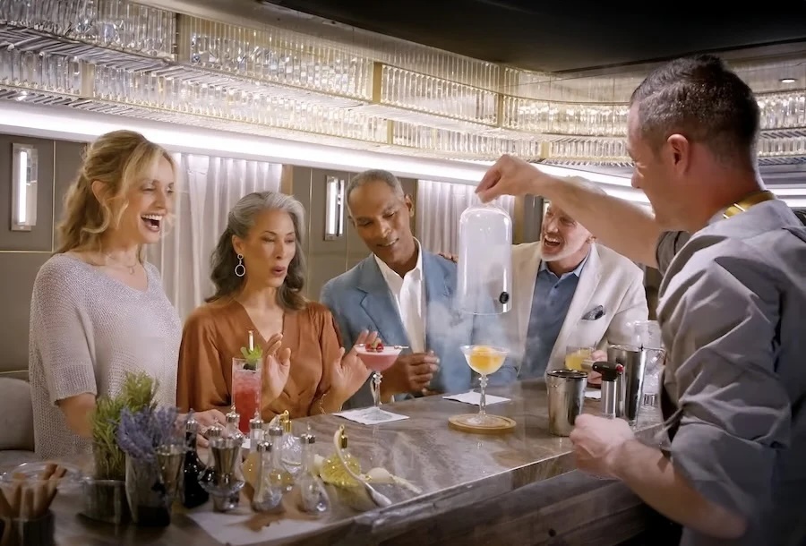 Bartender reveals smoked cocktail under glass dome to four delighted patrons at a bar