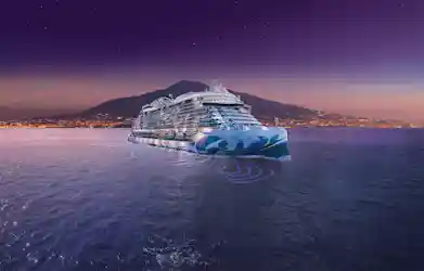 Illuminated Norwegian Viva cruise ship at dusk, sailing past coastal city with mountain and stars.