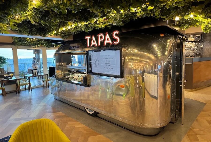 Silver Airstream-style tapas kiosk with neon 'TAPAS' sign under leafy ceiling in dining area.