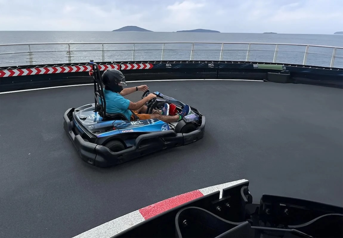Adult wearing helmet driving blue go-kart on cruise ship deck track with sea and islands.