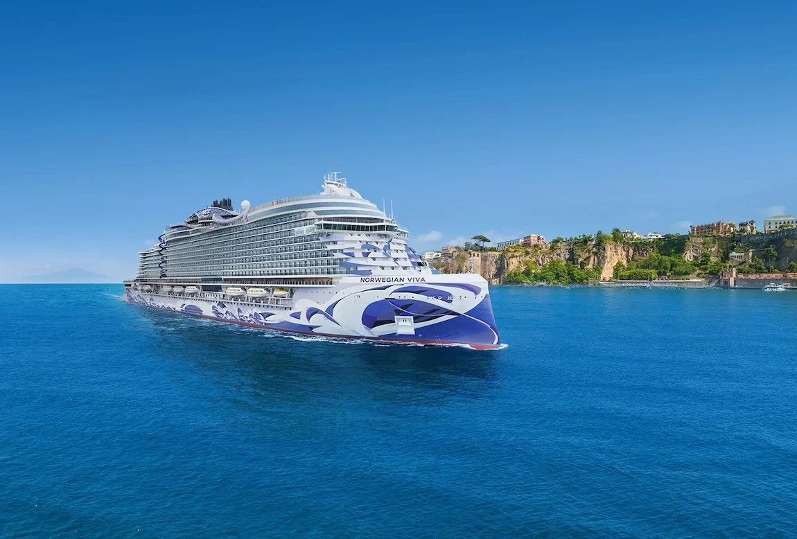 Large Norwegian Viva cruise ship sailing near sunlit coastal cliffs with seaside buildings.