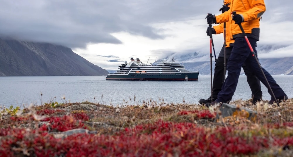 Two hikers in yellow jackets with poles on red tundra, expedition ship in foggy mountain fjord