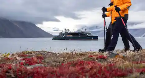 Two hikers in yellow jackets with poles on red tundra, expedition ship in foggy mountain fjord