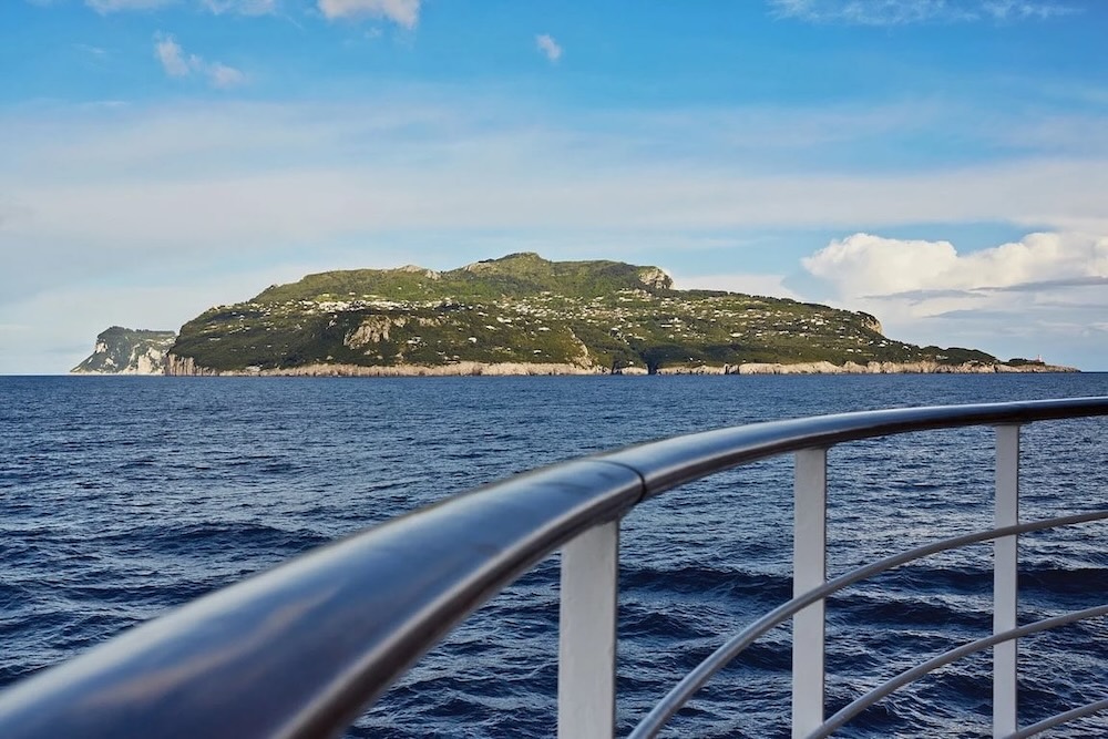 Ship railing foreground with blue sea and a distant green island under a bright sky