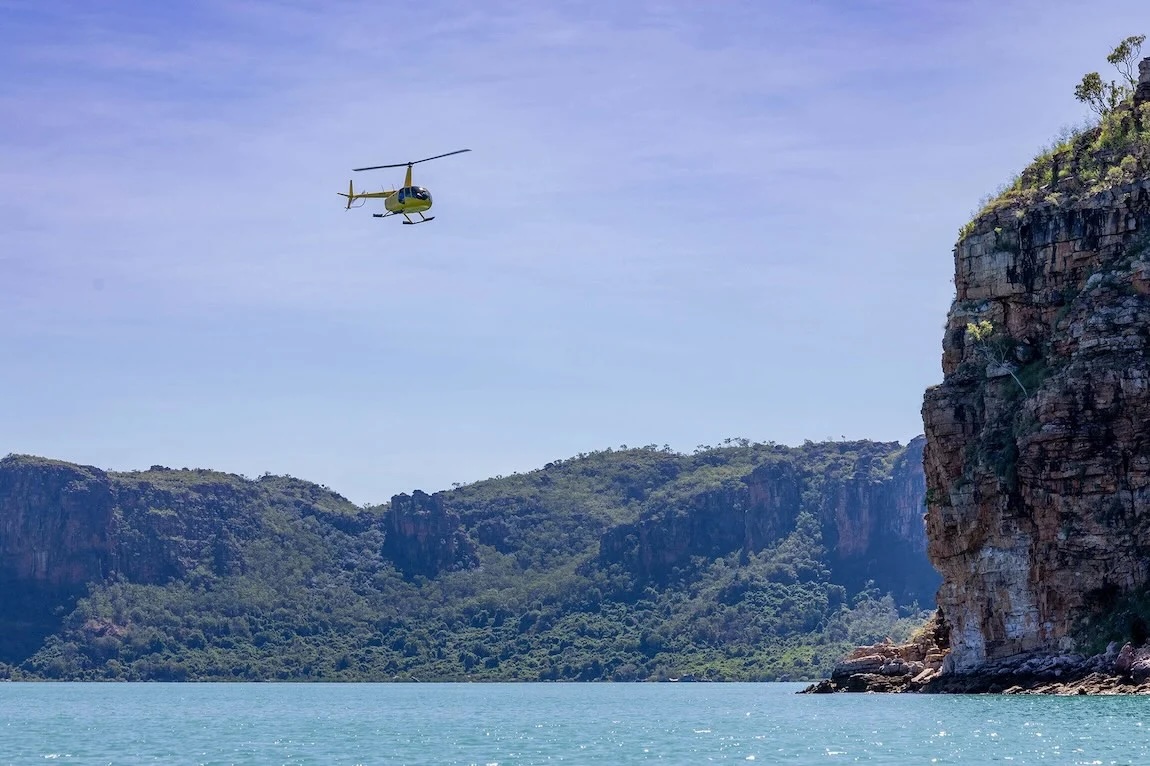 Yellow helicopter over turquoise water near forested cliffs and rocky headland.