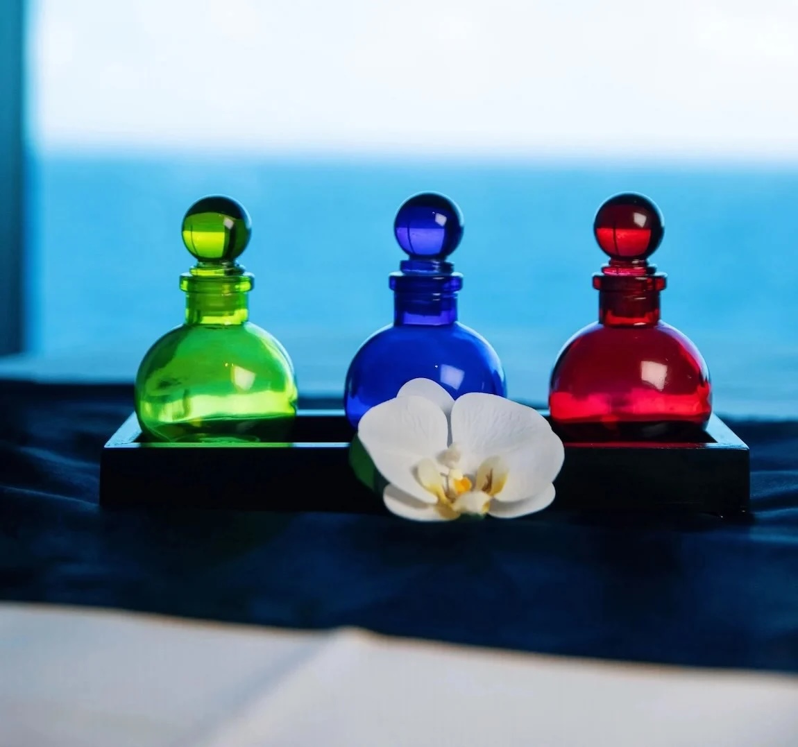 Three round glass bottles (green, blue, red) on black tray with white orchid, ocean view