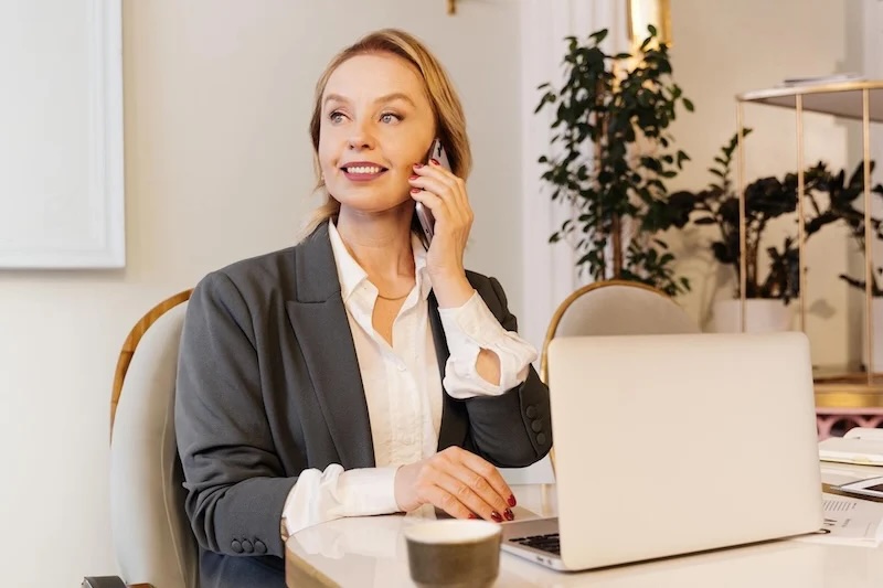 Businesswoman on phone at desk, smiling, laptop and coffee cup in modern office with plants