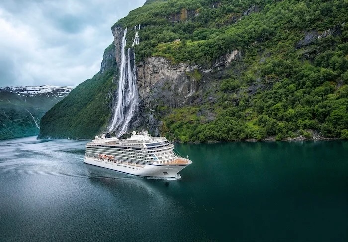 Cruise ship sailing a deep green fjord beneath tall cascading waterfall and steep cliffs