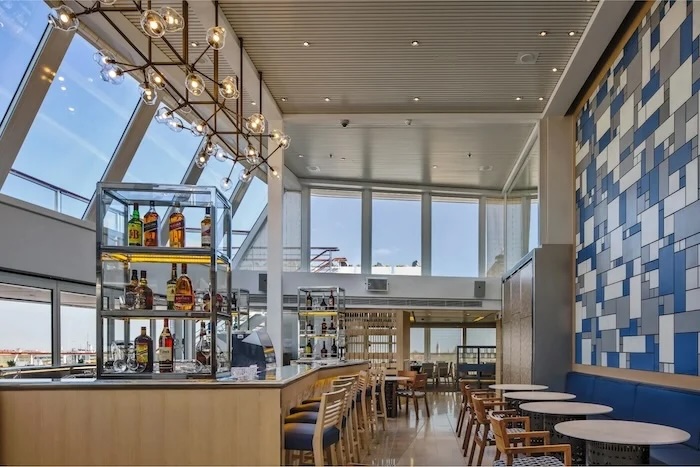 Modern airport lounge bar with backlit liquor display, bar stools, round tables, high windows.