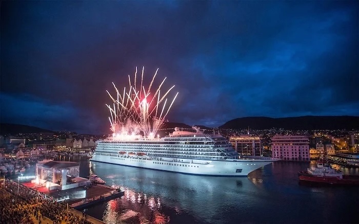 White cruise ship in harbor at night with fireworks overhead and celebrating crowd