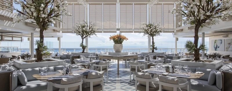 Bright seaside dining room with round tables, banquettes, potted trees and ocean view.