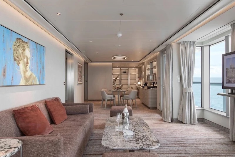 Ocean-view suite lounge with sofa, marble table with bottle and glasses, dining area
