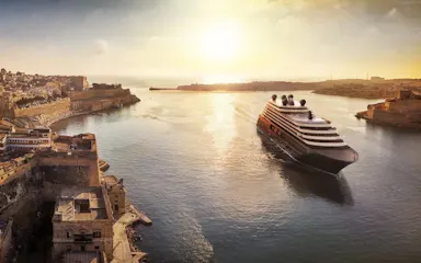 Large cruise ship entering sunlit harbor past historic fortified limestone city at sunset