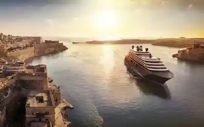 Large cruise ship entering sunlit harbor past historic fortified limestone city at sunset
