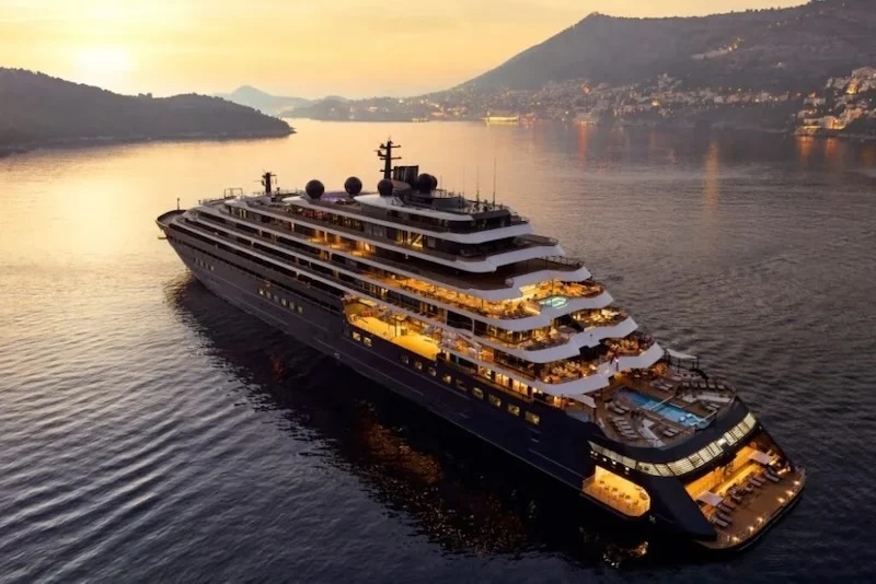 Luxury multi-deck cruise ship with illuminated decks and pools sailing near coastal hills at sunset