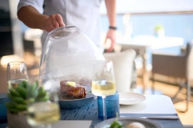 Hand lifts glass cloche to reveal butter-topped steak on plate at set dining table