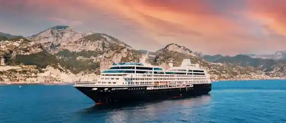 Passenger cruise ship sailing along a rocky Mediterranean coastline beneath a pink sunset sky