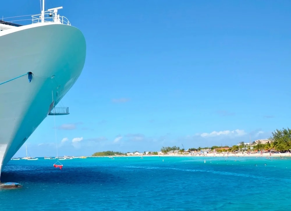 Docked cruise ship bow over turquoise sea with hanging lifeboat and crowded sandy beach