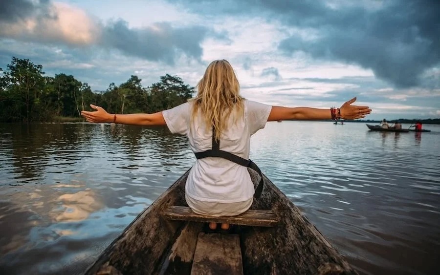 Person with outstretched arms seated in wooden canoe on a wide river, trees and boats ahead