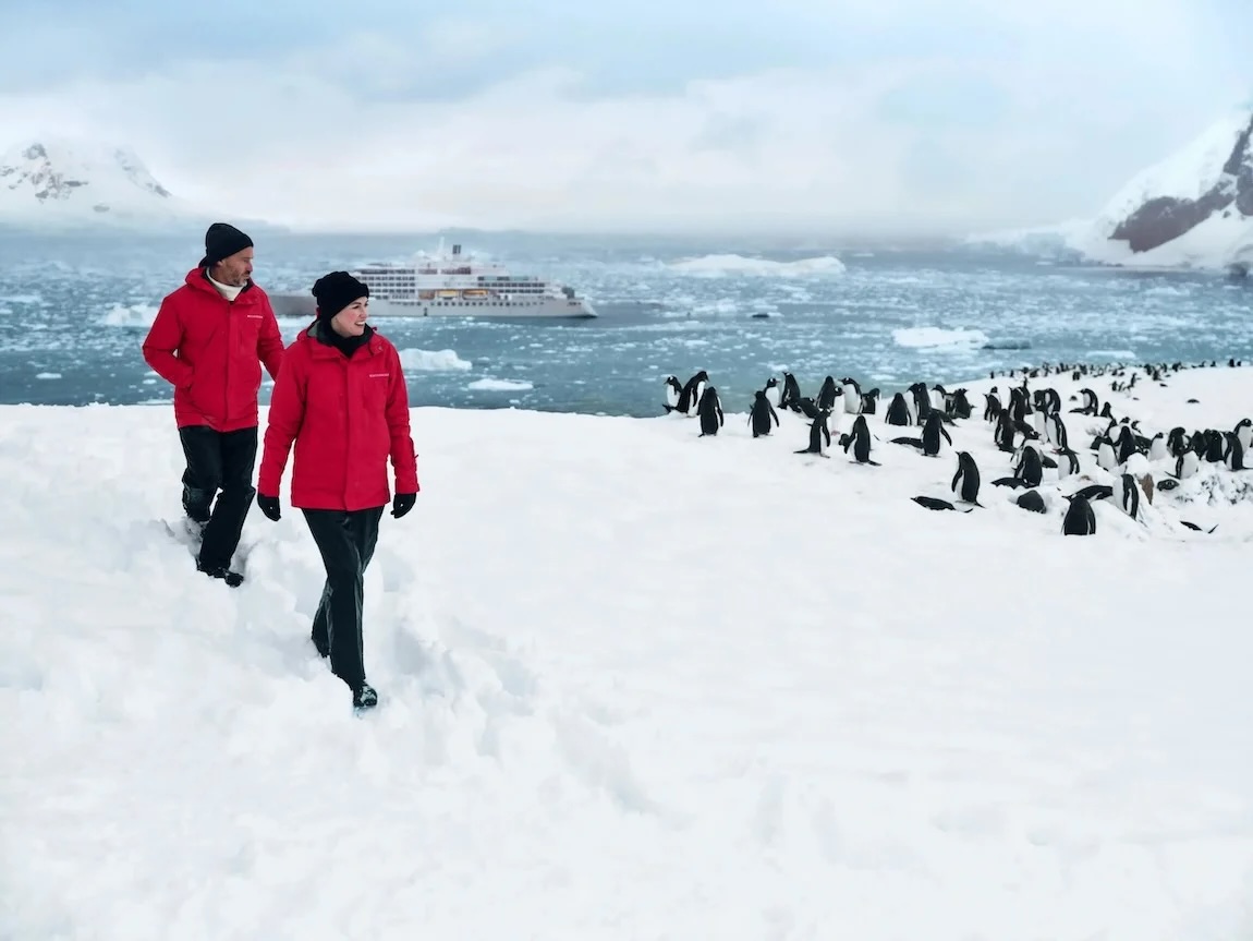 Two people in red parkas on snowy Antarctic shore beside penguin colony and cruise ship
