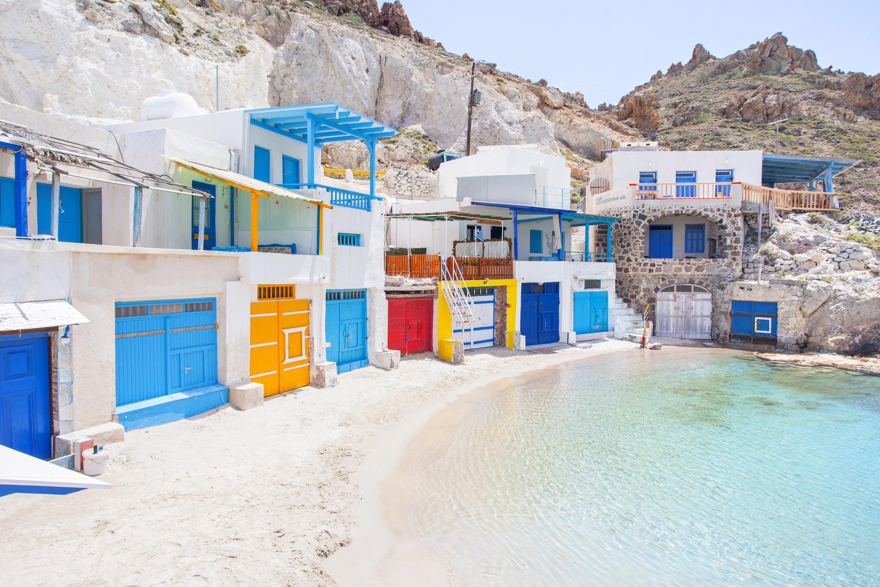 Whitewashed boathouses built into rocky cliffs with blue, yellow and red doors by turquoise water