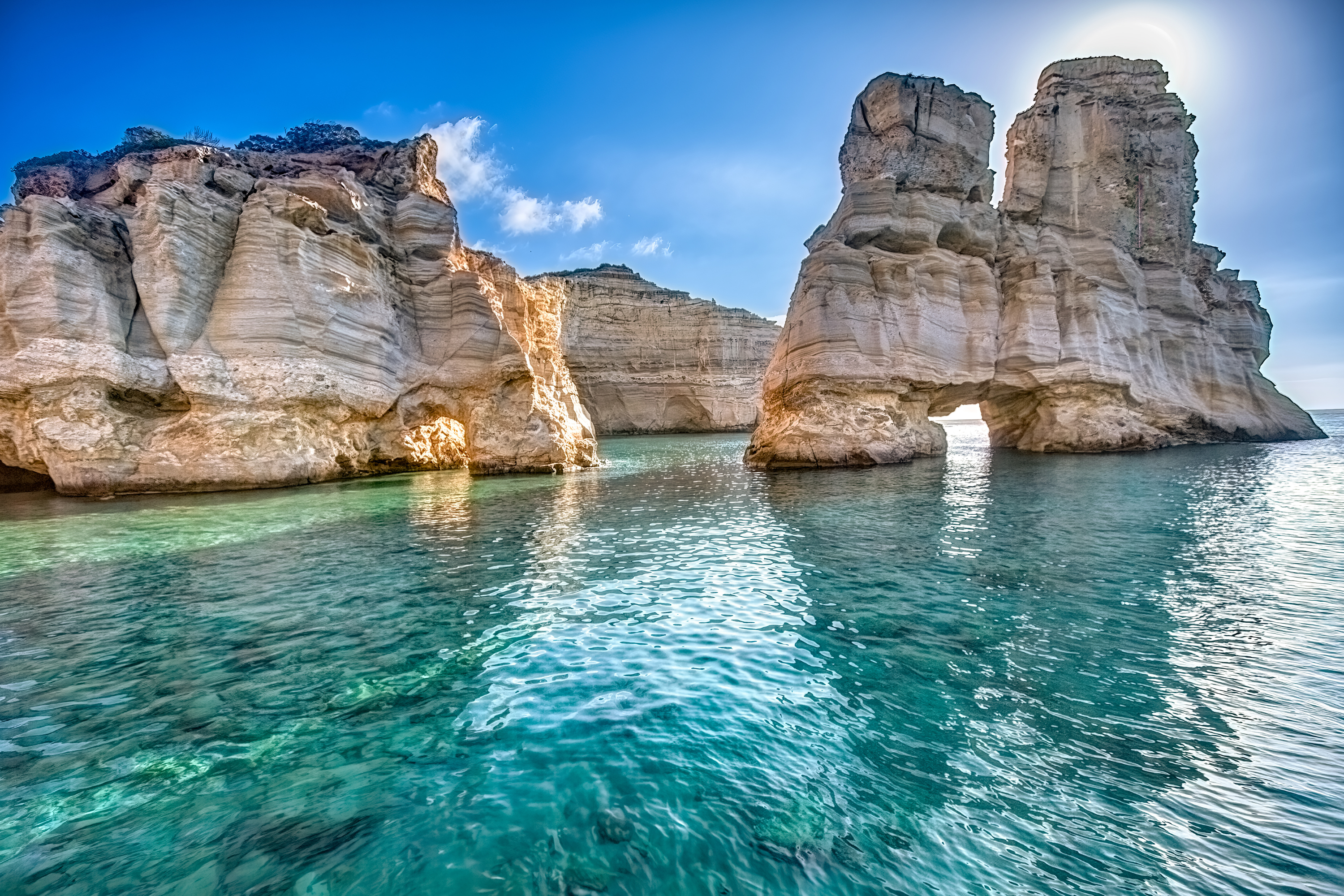 Turquoise sea with towering layered limestone cliffs and a sunlit natural rock arch.