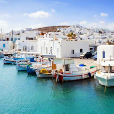 Fishing boats moored in turquoise harbor before whitewashed Greek island village and a Greek flag