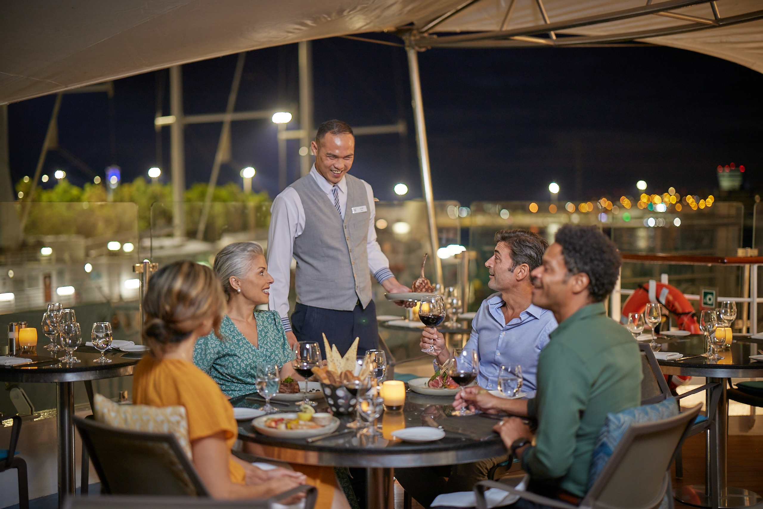 Smiling waiter serves steak to four people enjoying wine at an outdoor marina dinner