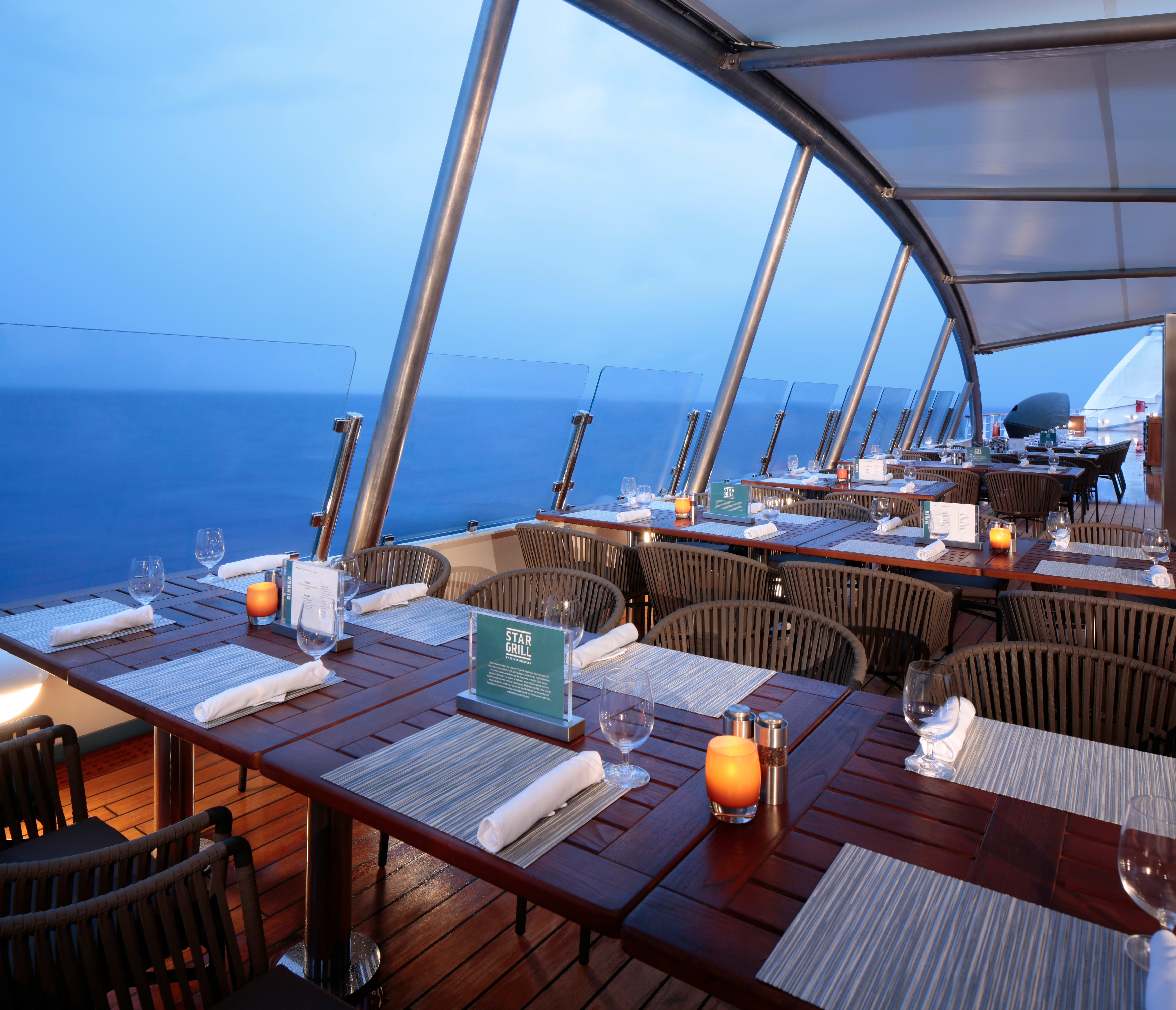 Cruise ship open-air dining deck with set tables, candles and glass railings overlooking the ocean.