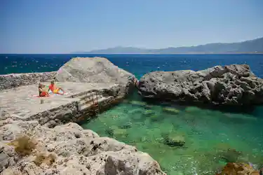 Two sunbathers on a stone pier beside a turquoise cove with ladder and distant mountains