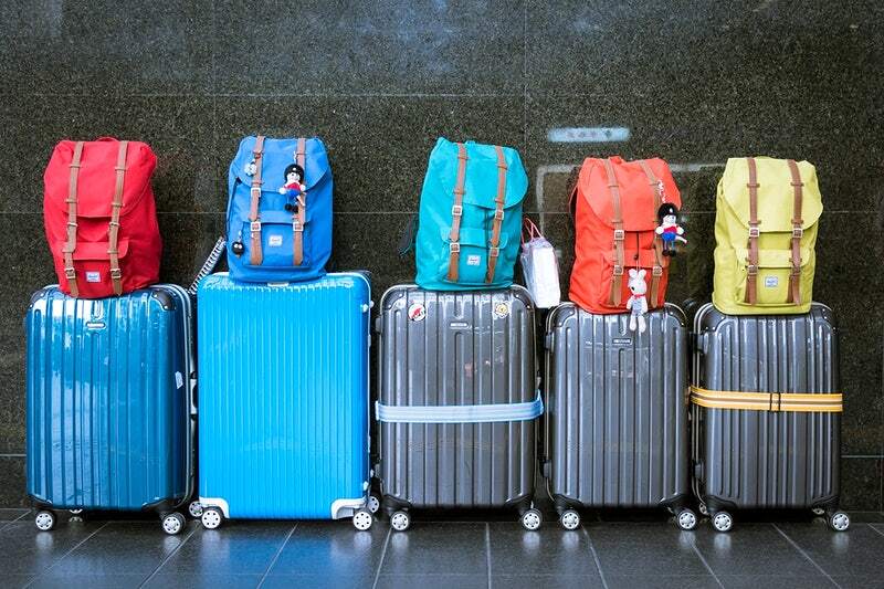 Row of six wheeled suitcases against dark wall, each with a colorful backpack and travel tags